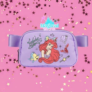 May include: A lavender fanny pack featuring Ariel from The Little Mermaid. The design includes Ariel, Flounder, Sebastian, and other themed graphics. The bag has a zip closure and a lavender strap.