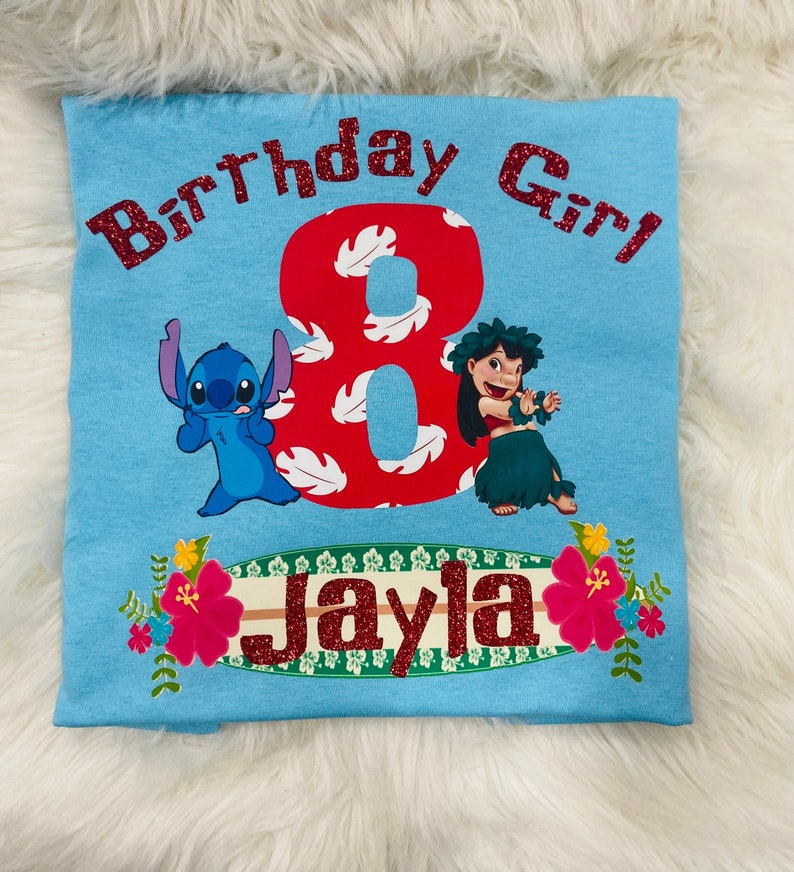 Peut inclure: T-shirt bleu clair avec un chiffre 8 rouge, un personnage bleu Stitch, une fille aux cheveux bruns en jupe hula verte, et le texte "Birthday Girl" et "Jayla" en paillettes rouges.