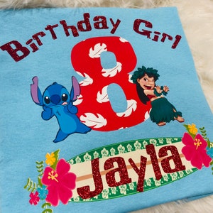 Peut inclure: T-shirt bleu clair avec un chiffre 8 rouge, des feuilles tropicales blanches et rouges, un personnage bleu Stitch, une fille aux cheveux bruns en jupe de hula verte, et le texte "Birthday Girl" et "Jayla" en paillettes rouges.