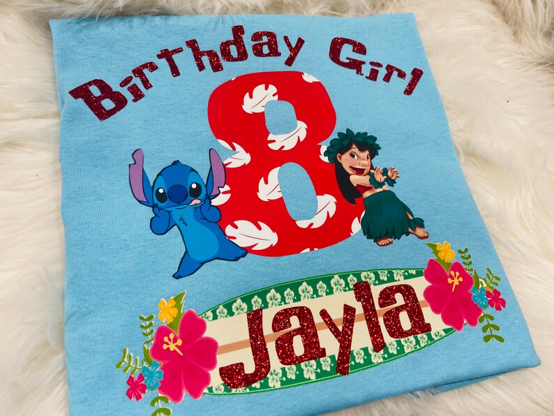 Peut inclure: T-shirt bleu clair avec un chiffre 8 rouge, des feuilles tropicales blanches et rouges, un personnage bleu Stitch, une fille aux cheveux bruns en jupe de hula verte, et le texte "Birthday Girl" et "Jayla" en paillettes rouges.
