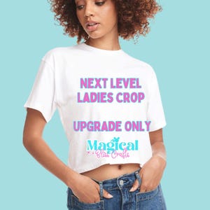 May include: White crop top with the text "NEXT LEVEL LADIES CROP UPGRADE ONLY Magical Bliss Crafts" in pink and blue. The model is wearing blue jeans and has curly brown hair.