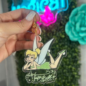 Puede incluir: Un llavero transparente de acrílico de Campanilla con un cierre dorado. El llavero presenta una ilustración de dibujos animados de Campanilla con un vestido verde, con las palabras "Tinker Bell" escritas en una fuente decorativa.