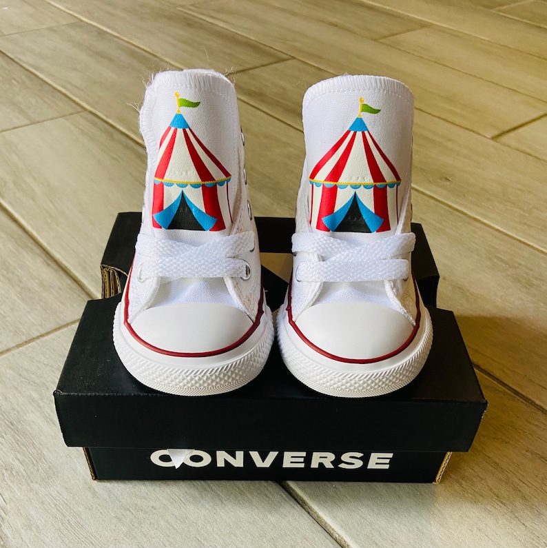 K&ouml;nnte beinhalten: Ein Paar wei&szlig;e Converse-Sneaker mit hohem Schaft, auf denen sich auf jedem Schuh ein rotes, wei&szlig;es und blaues Zirkuszelt befindet. Die Schuhe stehen auf einem schwarzen Kasten mit dem Aufdruck "CONVERSE".
