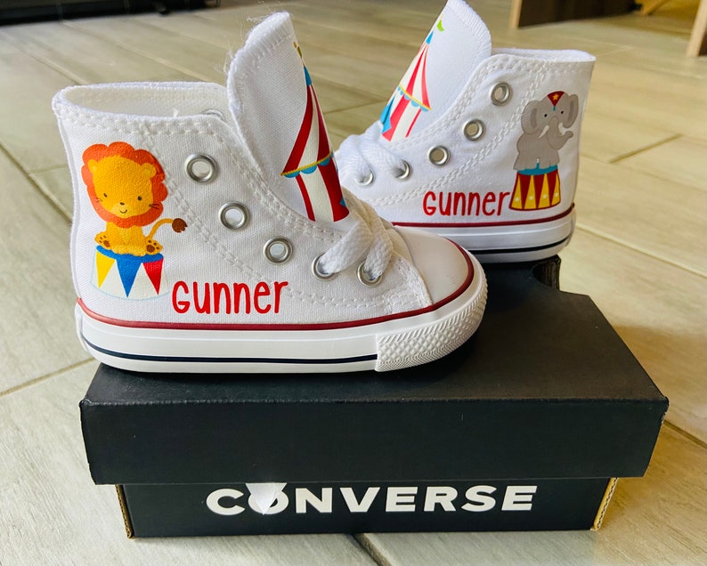K&ouml;nnte beinhalten: Wei&szlig;e Converse-Sneaker mit hohem Schaft und einem roten, gelben und blauen Zirkuszeltdesign. Ein Schuh hat einen L&ouml;wen und der andere einen Elefanten. Der Name "Gunner" ist auf beiden Schuhen geschrieben.