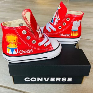 K&ouml;nnte beinhalten: Rote Converse-Sneaker mit hohem Schaft im Zirkus-Design. Ein Schuh zeigt einen L&ouml;wen, der andere einen Elefanten. Beide Schuhe tragen den Namen "Chase" auf sich.