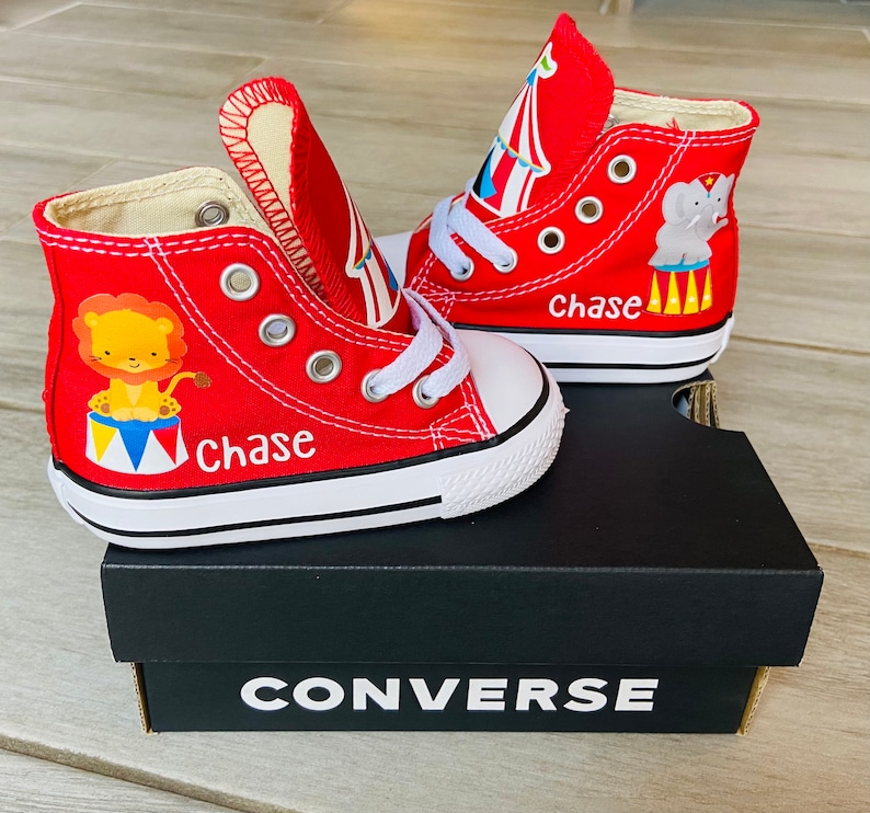 K&ouml;nnte beinhalten: Rote Converse-Sneaker mit hohem Schaft im Zirkus-Design. Ein Schuh zeigt einen L&ouml;wen, der andere einen Elefanten. Beide Schuhe tragen den Namen "Chase" auf sich.