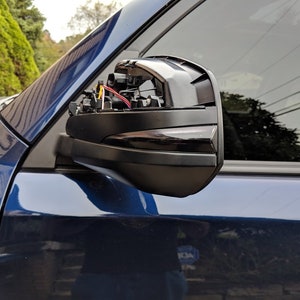 May include: A close-up of a black side mirror on a blue vehicle. The mirror is partially disassembled, revealing wires and internal components. The vehicle's window and body are visible in the background.