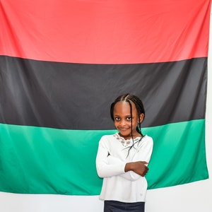 May include: A young girl with braids stands in front of a red, black, and green flag. She is wearing a white long-sleeved shirt and a dark skirt. She has her arms crossed and is smiling at the camera.