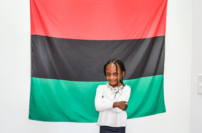 May include: A young girl with braids stands in front of a red, black, and green flag. She is wearing a white long-sleeved shirt and a dark skirt. She has her arms crossed and is smiling at the camera.