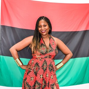 May include: A woman wearing a red and black patterned dress with gold bracelets stands in front of a red, black, and green flag.
