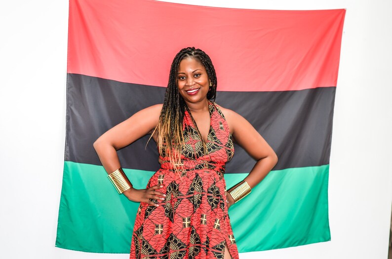 May include: A woman wearing a red and black patterned dress with gold bracelets stands in front of a red, black, and green flag.