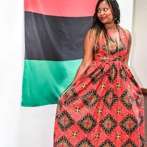 May include: A woman wearing a red, black, and gold patterned maxi dress stands in front of a red, black, and green flag. The dress has a halter neckline and a fitted bodice. The flag is hanging on a white wall.