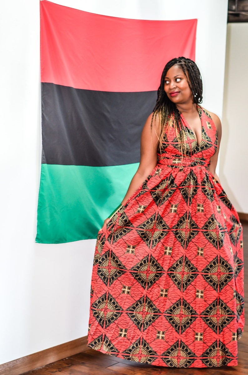 May include: A woman wearing a red, black, and gold patterned maxi dress stands in front of a red, black, and green flag. The dress has a halter neckline and a fitted bodice. The flag is hanging on a white wall.