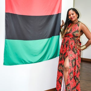 May include: A woman wearing a red, black, and gold patterned maxi dress with a side slit stands in front of a red, black, and green flag.