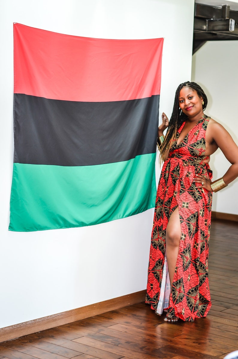 May include: A woman wearing a red, black, and gold patterned maxi dress with a side slit stands in front of a red, black, and green flag.