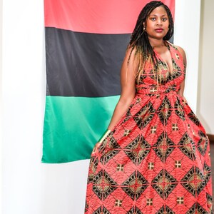 May include: A woman wearing a red, black, and gold patterned maxi dress stands in front of a red, black, and green flag.