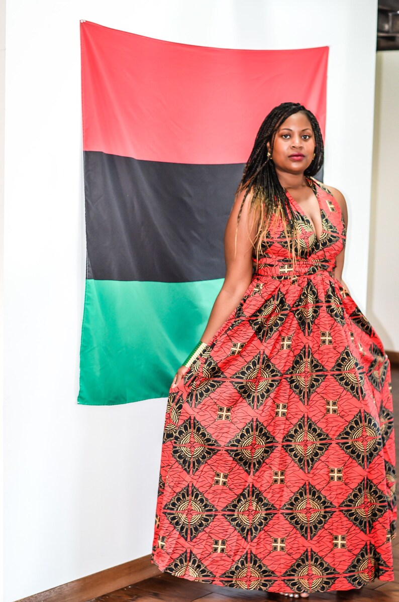 May include: A woman wearing a red, black, and gold patterned maxi dress stands in front of a red, black, and green flag.