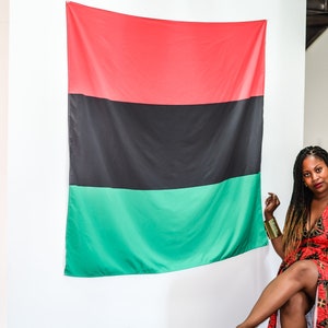 May include: A woman in a red and black patterned dress sits in front of a large red, black, and green flag. She is wearing gold bracelets on both wrists.