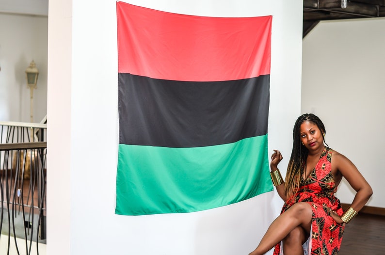 May include: A woman in a red and black patterned dress sits in front of a large red, black, and green flag. She is wearing gold bracelets on both wrists.