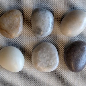 May include: Six smooth, rounded stones in various shades of gray, white, and brown. The stones are arranged in two rows of three on a textured, light brown surface.