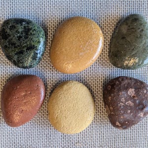 May include: Six smooth, rounded stones in various colors, including green, brown, yellow, and orange. The stones are arranged in two rows of three, on a textured, light gray surface.