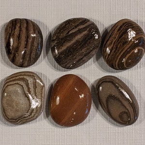 May include: Six smooth, oval-shaped stones with brown, gray, and white striped patterns. The stones have a glossy finish.