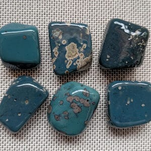 May include: Six polished gemstones in shades of teal and blue-green. The stones have rounded edges and a glossy finish, with some displaying mottled patterns of tan and cream. The arrangement is on a textured, neutral-toned surface.