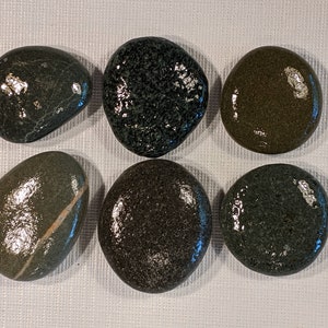 May include: Six smooth, polished stones in shades of green and gray. The stones vary in shape, with some oval and others more rounded. One stone has a white stripe. The stones are arranged on a white surface.