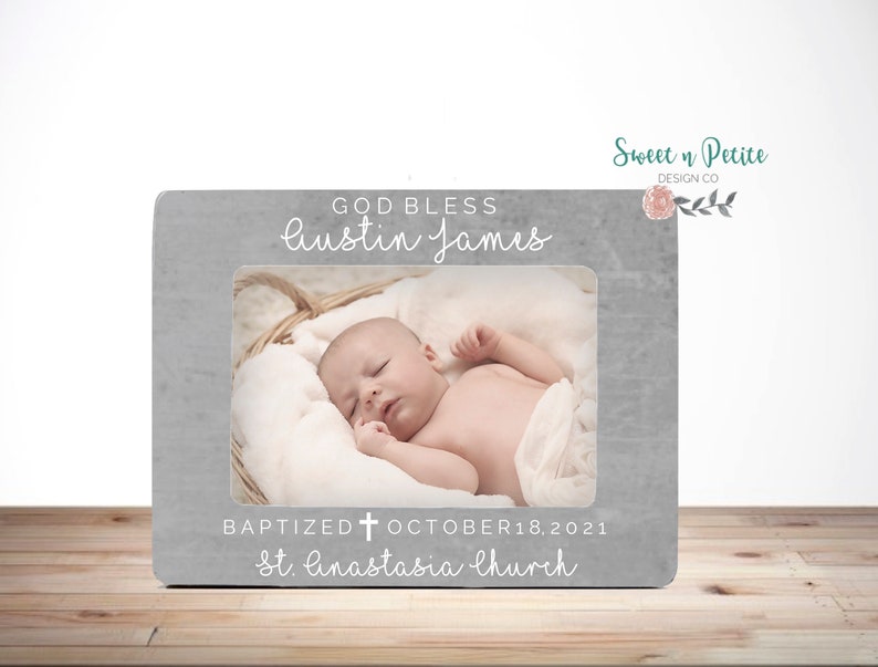 May include: A gray picture frame with a white border and a photo of a baby sleeping in a basket. The frame has the text "GOD BLESS Austin James BAPTIZED OCTOBER 18, 2021 St. Anastasia Church" on it.