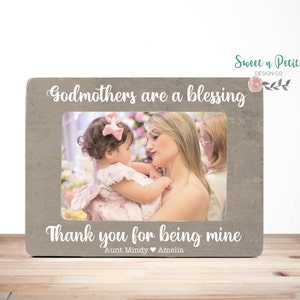 Könnte beinhalten: Ein rechteckiger, grauer Bilderrahmen mit dem Text "Godmothers are a blessing" und "Thank you for being mine." Der Rahmen enthält ein Foto einer Frau und eines Babys. Der Rahmen steht auf einer Holzoberfläche.