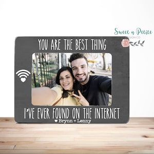 May include: A gray wooden picture frame with a white wifi symbol and a heart. The frame has a photo of a couple taking a selfie. The text on the frame reads "You are the best thing I've ever found on the internet" and "Brynn + Lenny".