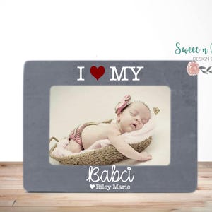 Puede incluir: Un marco de fotos gris con las palabras "I ❤️ MY Babci" y el nombre "Riley Marie" en escritura blanca. El marco contiene una foto de un bebé durmiendo en una cesta tejida.