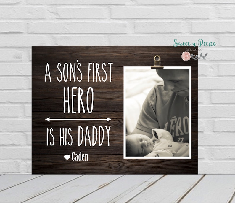May include: A wood-grain background with a white inscription that reads "A son's first hero is his daddy" with a heart and the name "Caden" below. A black and white photo is clipped to the background with a clothespin. The photo shows a father holding a baby.