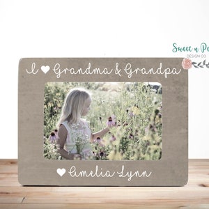 Op de afbeelding: Een bruin houten fotolijstje met een wit hartje en de tekst "Ik hou van oma & opa" bovenaan. Het lijstje heeft een foto van een jong meisje in een bloemenveld. De tekst "Amelia Lynn" staat onderaan het lijstje.