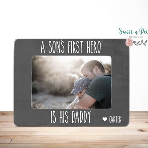 May include: A gray picture frame with the text "A son's first hero is his daddy" and a heart with the name "Carter". The frame has a photo of a father and son.
