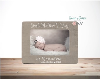 May include: A light brown wooden picture frame with a white background and the text "First Mother's Day as Grandma Love, Sophia 2022". The frame has a photo of a baby girl sleeping in it.