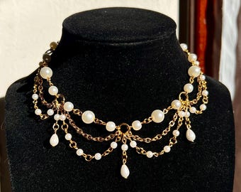 Puede incluir: Un collar dorado con múltiples hilos de pequeñas perlas blancas y perlas redondas más grandes. El collar tiene un diseño delicado en cascada con colgantes de perlas en forma de lágrima. El collar se exhibe en un maniquí negro.