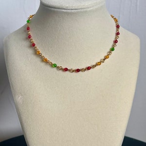 May include: A gold-colored beaded necklace with red, yellow, and green beads. The necklace is displayed on a white mannequin bust against a white background. The beads are small and evenly spaced along the gold chain.