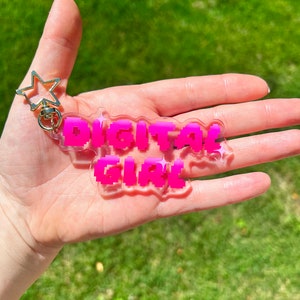 以下が含まれることがあります： 明るいピンク色のピクセル化された文字で「DIGITAL GIRL」と書かれた透明なアクリルキーホルダー。キーホルダーには、金色の星型の留め金と金色のリングが付いています。背景は緑の草です。