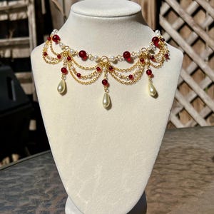 Peut inclure: Un collier doré orné de perles rouges, de perles blanches et de pendentifs en forme de goutte. Le collier est présenté sur un buste de mannequin blanc. Le design comprend plusieurs rangées de chaînes fines, probablement des bijoux fantaisie.