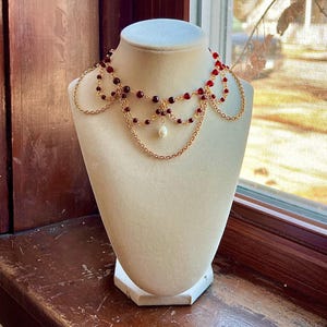 May include: A gold-toned necklace with cascading chains and red beads. The necklace features a central pearl pendant and is displayed on a cream-colored mannequin. The design is intricate and elegant, suitable for special occasions.