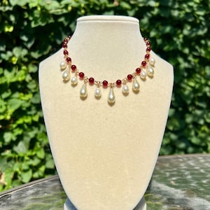 May include: A gold necklace featuring red beads and hanging white pear-shaped pearls. The necklace is displayed on a cream mannequin against a backdrop of green leaves.