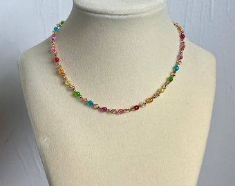 May include: A colorful beaded necklace displayed on a white mannequin. The necklace features small, round beads in various colors, strung together with gold-colored chain. The beads include shades of red, pink, yellow, green, and blue.