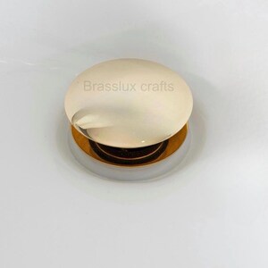 May include: A round, gold-toned sink stopper with a polished finish. The stopper has a circular, off-white top with the text "Brasslux crafts" printed on it. The base is a contrasting silver color.