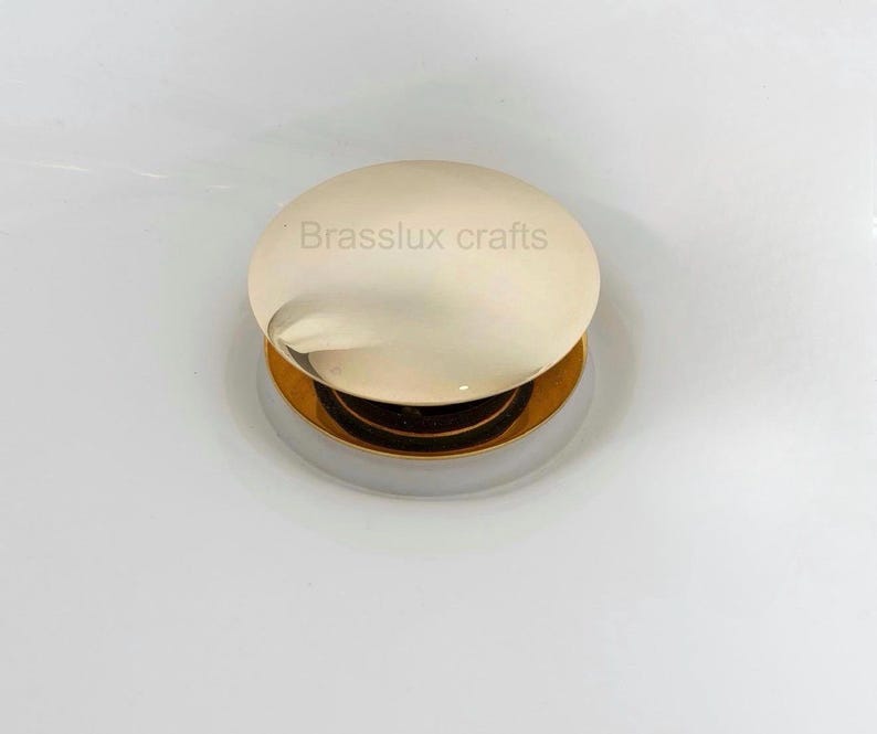 May include: A round, gold-toned sink stopper with a polished finish. The stopper has a circular, off-white top with the text "Brasslux crafts" printed on it. The base is a contrasting silver color.