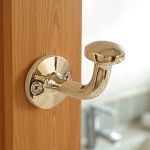 May include: A polished brass coat hook mounted on a wooden surface. The hook features a rounded base with two screws and a curved arm ending in a circular knob. The finish is shiny and reflective.