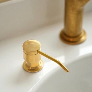 May include: A close-up of a gold-colored soap dispenser with a curved spout. The dispenser is cylindrical with a round base and a pump top. The background is a white sink and a gold faucet.