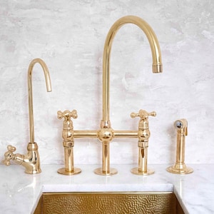 May include: A set of four gold-colored kitchen faucets. The set includes a tall, arched faucet, two faucets with cross-shaped handles, and a side sprayer. The faucets are mounted on a white countertop.