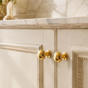 May include: Two polished gold-toned round cabinet knobs with a tiered, circular design attached to off-white, paneled wooden cabinet doors. The knobs feature a smooth, reflective surface and a central stem connecting to the cabinet face.