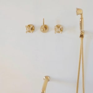 Pode incluir: Torneiras douradas para casa de banho contra uma parede branca. O conjunto inclui um chuveiro com mangueira flex&iacute;vel, um bico curvo e tr&ecirc;s comandos de parede. Os acess&oacute;rios t&ecirc;m um design cl&aacute;ssico e elegante.